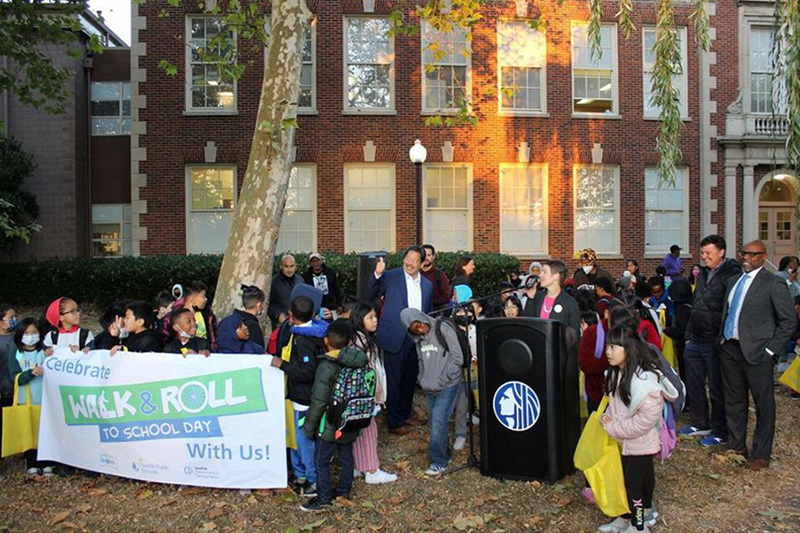 Seattle Mayor Bruce Harrell gives a thumbs up and Nancy Pullen-Seufert shares her remarks while students gather around the podium at Dunlap Elementary School in Rainier Beach. Photo credit: Seattle Mayor's Office.